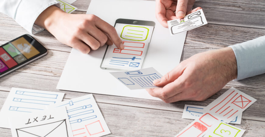 Close up of two designers holding pieces of cut paper with sketches of website layouts above a print-out of a smartphone on a light gray wood desk.