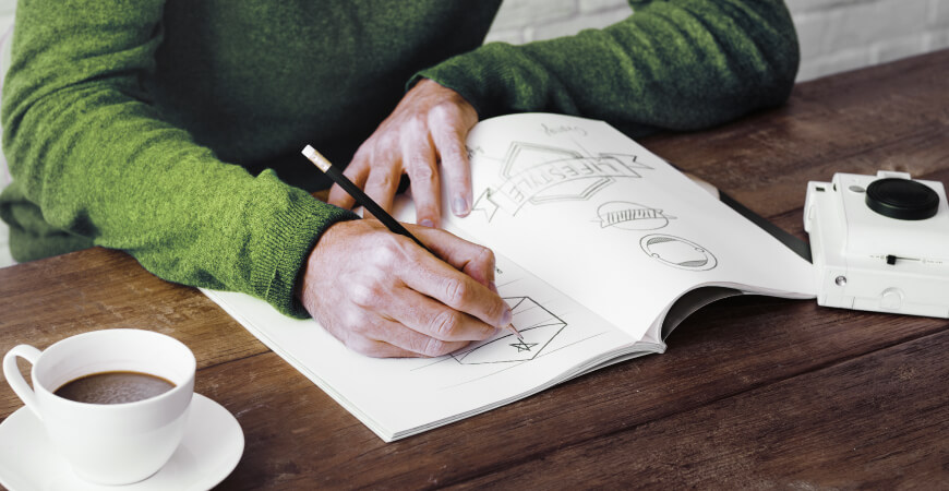 Close up of a male graphic designer wearing a green sweater sketching logo ideas on a notepad on a dark wooden table with a cup of coffee and small white camera.